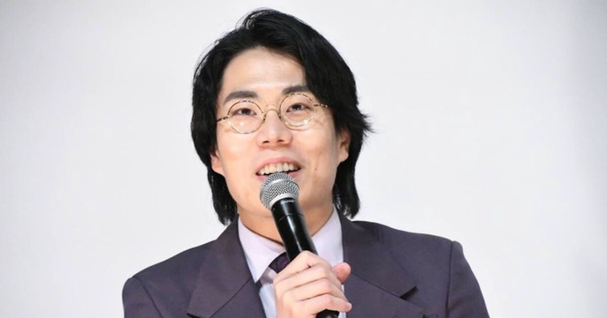 黒髪と丸メガネを身につけたスーツ姿の人物がマイクを持って話している白い背景の画像
